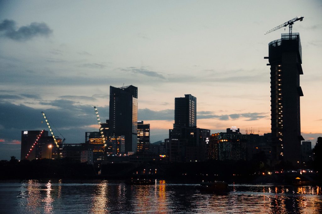 Evening skyline with skyscrapers and cranes reflecting on a river, showcasing a city under construction at sunset.